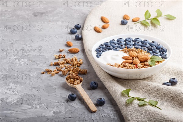 Yoghurt with blueberry, granola and almond in white plate with wooden spoon on gray concrete background and linen textile. side view, copy space