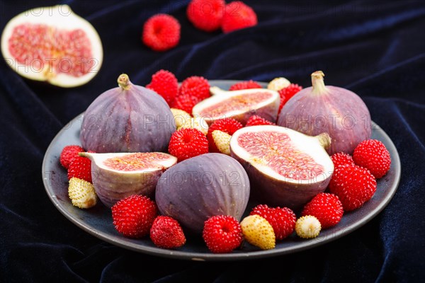 Fresh figs, strawberries and raspberries on blue ceramic plate on black concrete background and blue velvet textile. side view, close up, selective focus