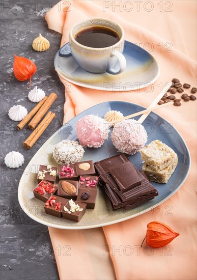 A pieces of homemade chocolate with coconut candies and a cup of coffee on a black concrete background and orange textile. side view, close up