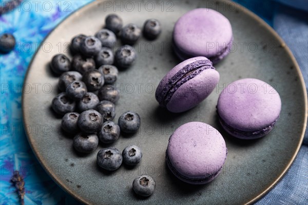 Purple macarons or macaroons cakes with blueberries on ceramic plate on a blue concrete background and blue textile. Side view, close up, selective focus