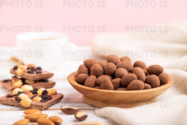Almond in chocolate dragees in wooden plate and a cup of coffee on white and pink background and linen textile. Side view, copy space, selective focus
