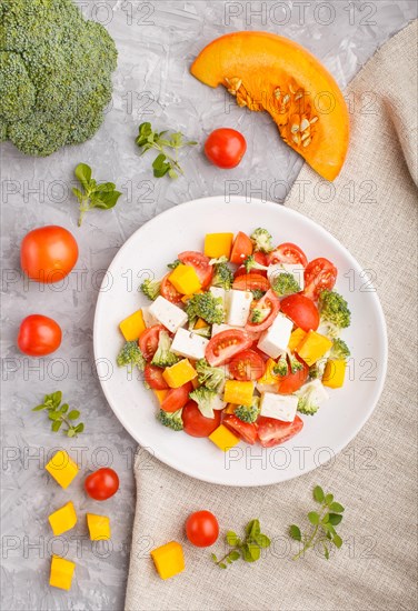 Vegetarian salad with broccoli, tomatoes, feta cheese, and pumpkin on white ceramic plate on a gray concrete background and linen textile, top view, close up, flat lay