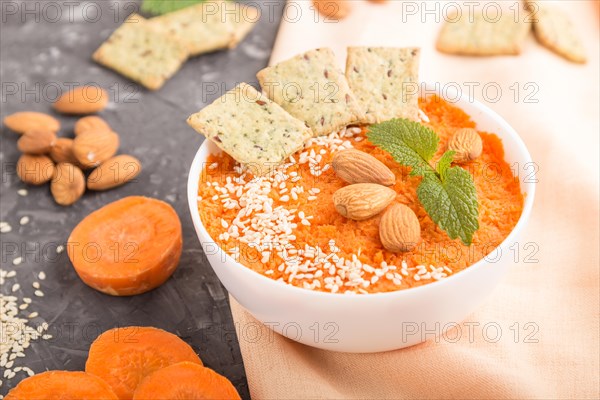 Carrot cream soup with sesame seeds, almonds and snacks in white bowl on a black concrete background with orange textile. side view, close up, selective focus