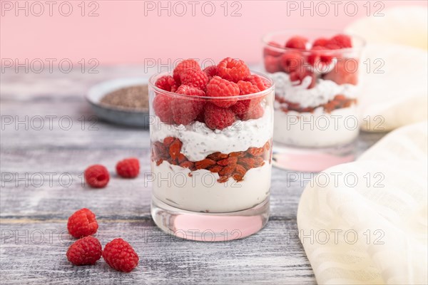 Yogurt with raspberry, goji berries and chia seeds in glass on gray wooden background and linen textile. Side view, close up, selective focus