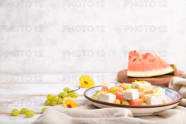 Vegetarian salad with watermelon, feta cheese, and grapes on blue ceramic plate on white wooden background and linen textile. Side view, copy space, selective focus