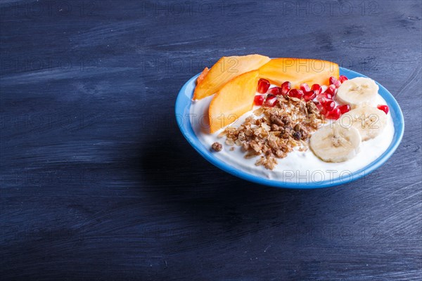 A blue plate with greek yogurt, granola, persimmon, banana, pomegranate black wooden background. close up. copy space