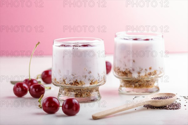 Yoghurt with cherries, chia seeds and granola in glass with wooden spoon on pink and white background. side view, close up, selective focus, copy space