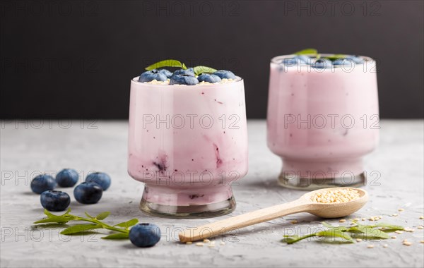 Yoghurt with blueberry and sesame in a glass and wooden spoon on gray and black background. side view, close up