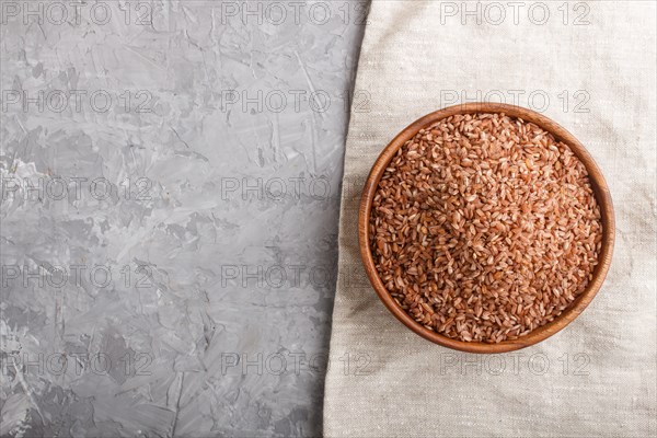 Wooden bowl with unpolished brown rice on a gray concrete background and linen textile. Top view, flat lay, copy space
