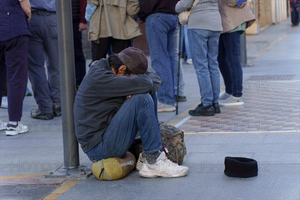Homeless man on the street begging for alms with a sad expression, his head between his legs and a cap on the ground for coins
