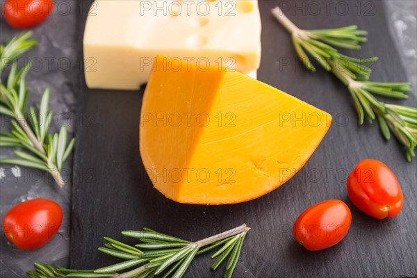 Various types of cheese with rosemary and tomatoes on black slate board on a black concrete background. Side view, close up, selective focus