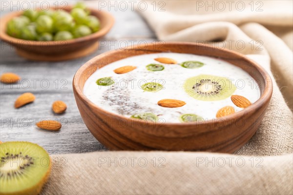 Yogurt with kiwi, gooseberry, chia and almonds in wooden bowl on gray wooden background and linen textile. Side view, close up, selective focus