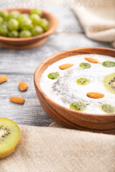 Yogurt with kiwi, gooseberry, chia and almonds in wooden bowl on gray wooden background and linen textile. Side view, close up, selective focus