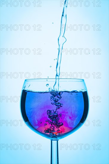 Pouring water into wine glass with red and blue liquid, illuminated in blue
