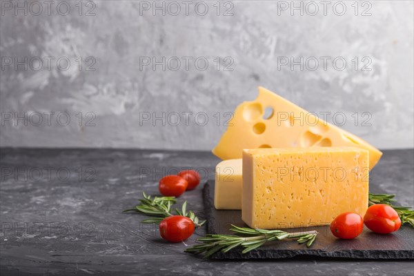 Various types of cheese with rosemary and tomatoes on black slate board on a black concrete background. Side view, close up, copy space, selective focus