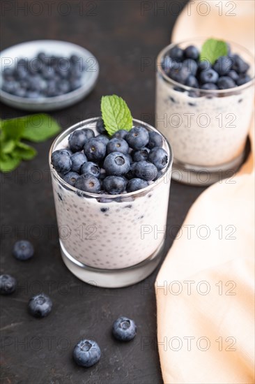Yogurt with blueberry and chia in glass on black concrete background and orange linen textile. Side view, close up