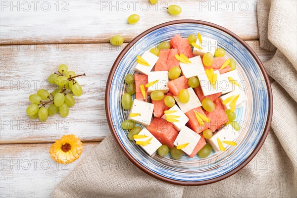 Vegetarian salad with watermelon, feta cheese, and grapes on blue ceramic plate on white wooden background and linen textile. top view, close up, flat lay