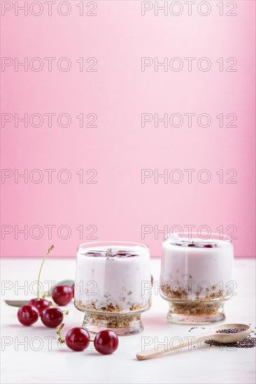 Yoghurt with cherries, chia seeds and granola in glass with wooden spoon on pink and white background. side view, close up, selective focus, copy space