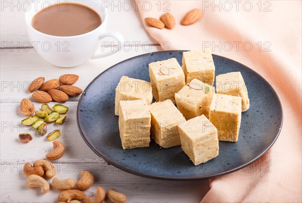 Traditional indian candy soan papdi in a blue ceramic plate with almond, pistache, cashew and a cup of coffee on a white wooden background with orange textile. side view, close up, selective focus