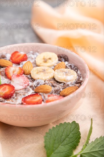 Chocolate cornflakes with milk, strawberry and almonds in ceramic bowl on gray concrete background and orange linen textile. Side view, close up, selective focus