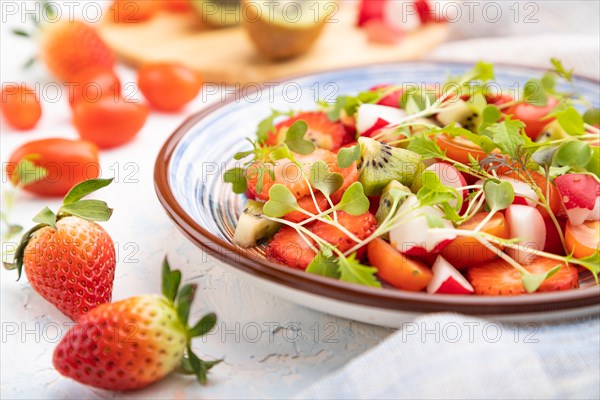 Vegetarian fruits and vegetables salad of strawberry, kiwi, tomatoes, microgreen sprouts on white concrete background and linen textile. Side view, close up, selective focus