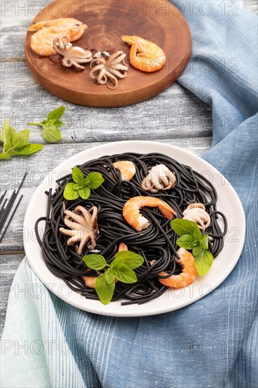 Black cuttlefish ink pasta with shrimps or prawns and small octopuses on gray wooden background and blue textile. Side view, close up