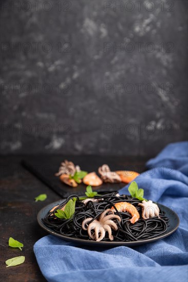 Black cuttlefish ink pasta with shrimps or prawns and small octopuses on black concrete background and blue textile. Side view, copy space, selective focus