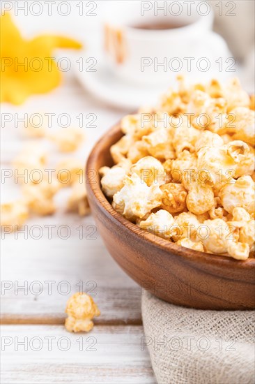 Popcorn with caramel in wooden bowl and a cup of coffee on a white wooden background and linen textile. Side view, close up, selective focus