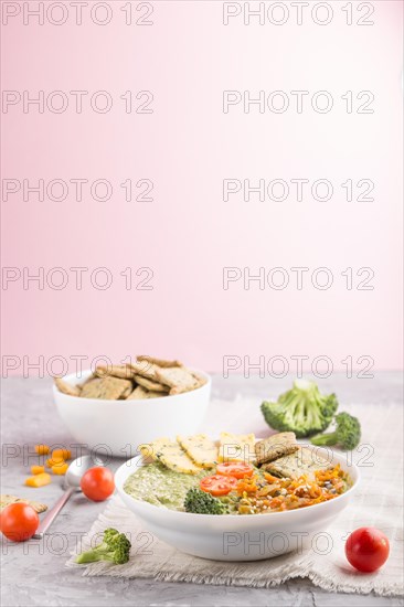 Green broccoli cream soup with crackers and cheese in white bowl on a gray and pink background and linen napkin. side view, copy space