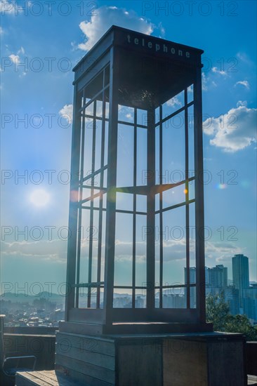 Red wooden telephone booth minus phone and without glass on roof of building back lit by the sun with blue sky and clouds in background in South Korea