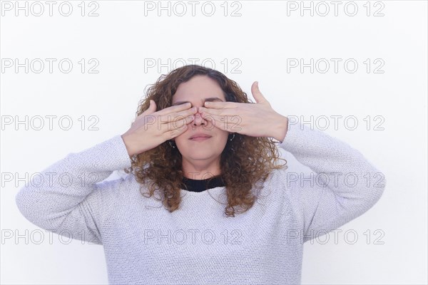 Beautiful blonde curly-haired woman dressed in white, covering her eyes with her hands on a white background and copyspace
