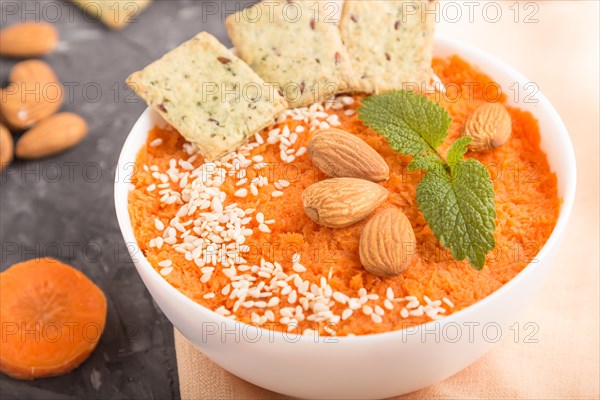 Carrot cream soup with sesame seeds, almonds and snacks in white bowl on a black concrete background with orange textile. side view, close up, selective focus