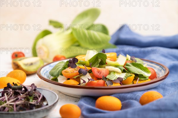 Vegetarian salad of pac choi cabbage, kiwi, tomatoes, kumquat, microgreen sprouts on a white concrete background and blue linen textile. Side view, close up, selective focus