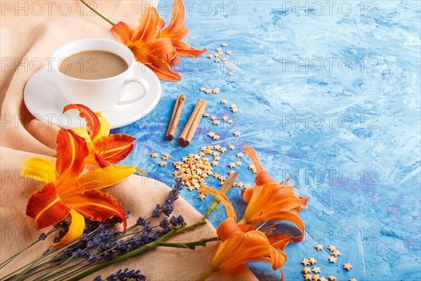 Orange day-lily and lavender flowers and a cup of coffee on a blue concrete background, with orange textile. Morninig, spring, fashion composition. side view, copy space