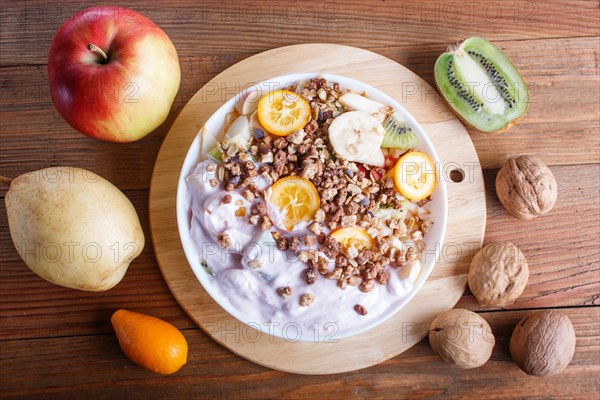 Vegetarian salad of bananas, apples, pears, kumquats, kiwi with granola and yogurt on brown woodem background, close up, top view