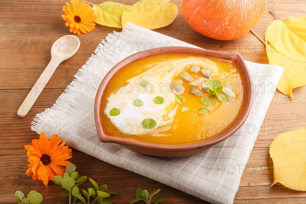 Traditional pumpkin cream soup with seeds in clay bowl on a brown wooden background with linen napkin. side view, close up