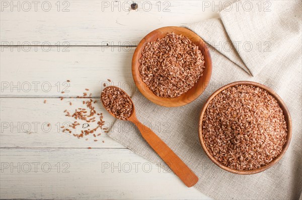 Two wooden bowls with unpolished brown rice and wooden spoon on a white wooden background and linen textile. Top view, flat lay, copy space