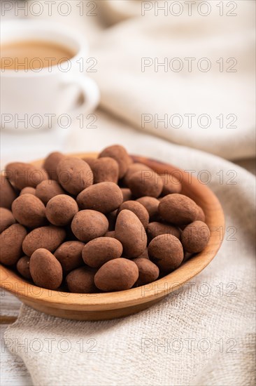 Almond in chocolate dragees in wooden plate and a cup of coffee on white wooden background and linen textile. Side view, close up, selective focus