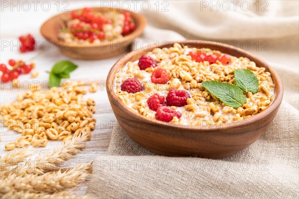 Wheat flakes porridge with milk, raspberry and currant in wooden bowl on white wooden background and linen textile. Side view, close up, selective focus
