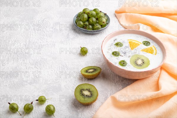 Yogurt with kiwi, gooseberry, chia in ceramic bowl on gray concrete background and orange linen textile. Side view, copy space