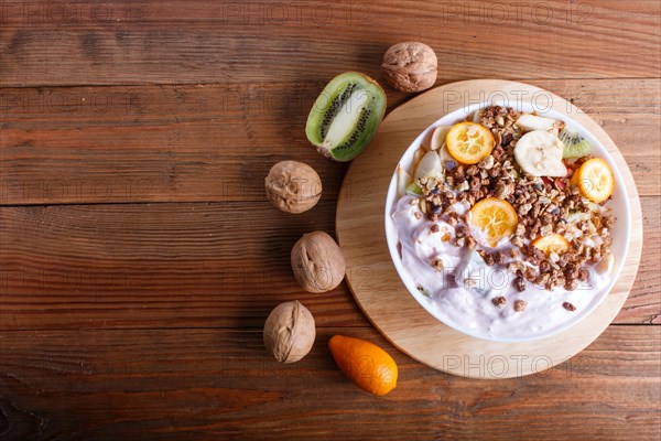 Vegetarian salad of bananas, apples, pears, kumquats, kiwi with granola and yogurt on brown woodem background, copy space, top view