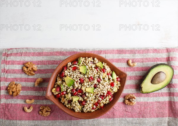 Salad of germinated buckwheat, avocado, walnut and pomegranate seeds in clay plate on white wooden background. Top view, copy space