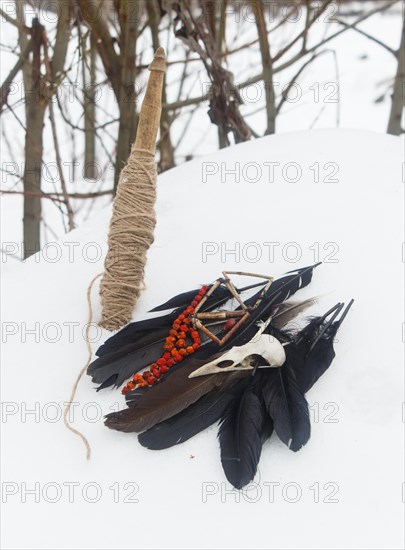 Spindle, crow feathers, bird skull and rowan beads in the snow. Still life. Pagan, witchcraft symbols