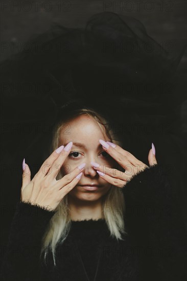 An intense muted close-up of a blonde caucasian young woman in dark lace clothing and hat, covering her eyes with her hands