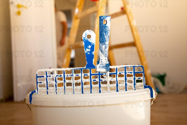 Symbolic image of painting work: Close-up of a paint bucket with scraper grid, in the background a painter's ladder