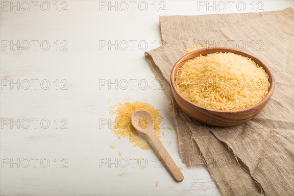Wooden bowl with raw golden rice and wooden spoon on a white wooden background and linen textile. Side view, close up, copy space