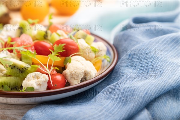 Vegetarian salad of cauliflower cabbage, kiwi, tomatoes, microgreen sprouts on white wooden background and blue linen textile. Side view, close up, selective focus