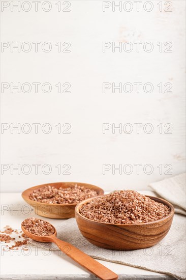 Two wooden bowls with unpolished brown rice and wooden spoon on a white wooden background and linen textile. Side view, copy space, vertical