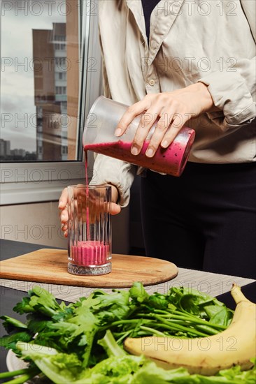Unrecognizable woman pouring banana and black currant smoothie in glass in the kitchen