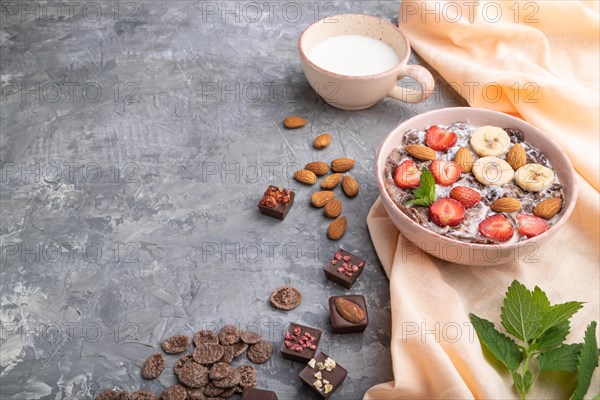 Chocolate cornflakes with milk, strawberry and almonds in ceramic bowl on gray concrete background and orange linen textile. Side view, copy space